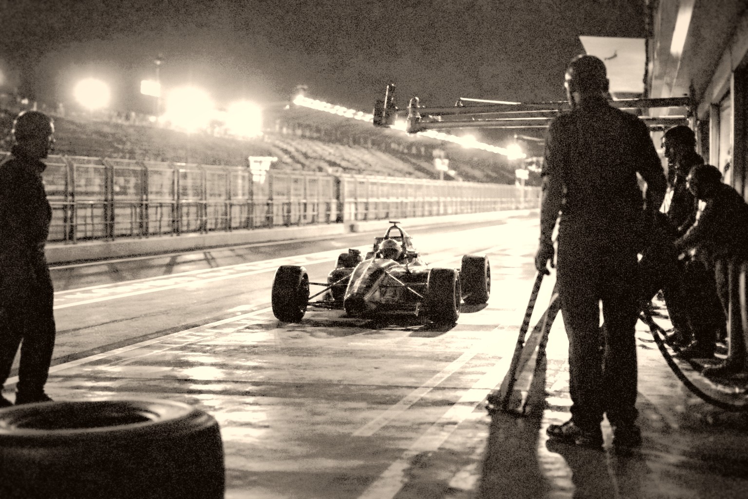 Pit lane nocturno con focos duros y mecánicos en silueta