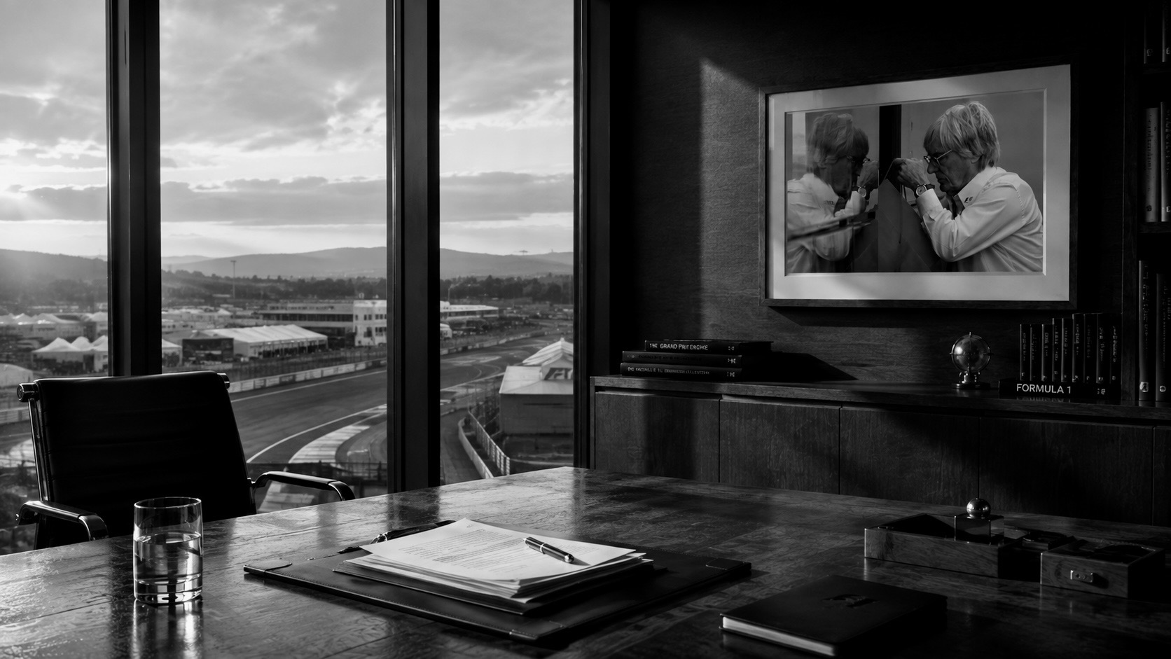 Despacho de dirección vacío al amanecer sobre un circuito, con una silla desplazada, un dossier abierto con una pluma encima, un vaso de agua y una fotografía enmarcada de Bernie Ecclestone en la pared