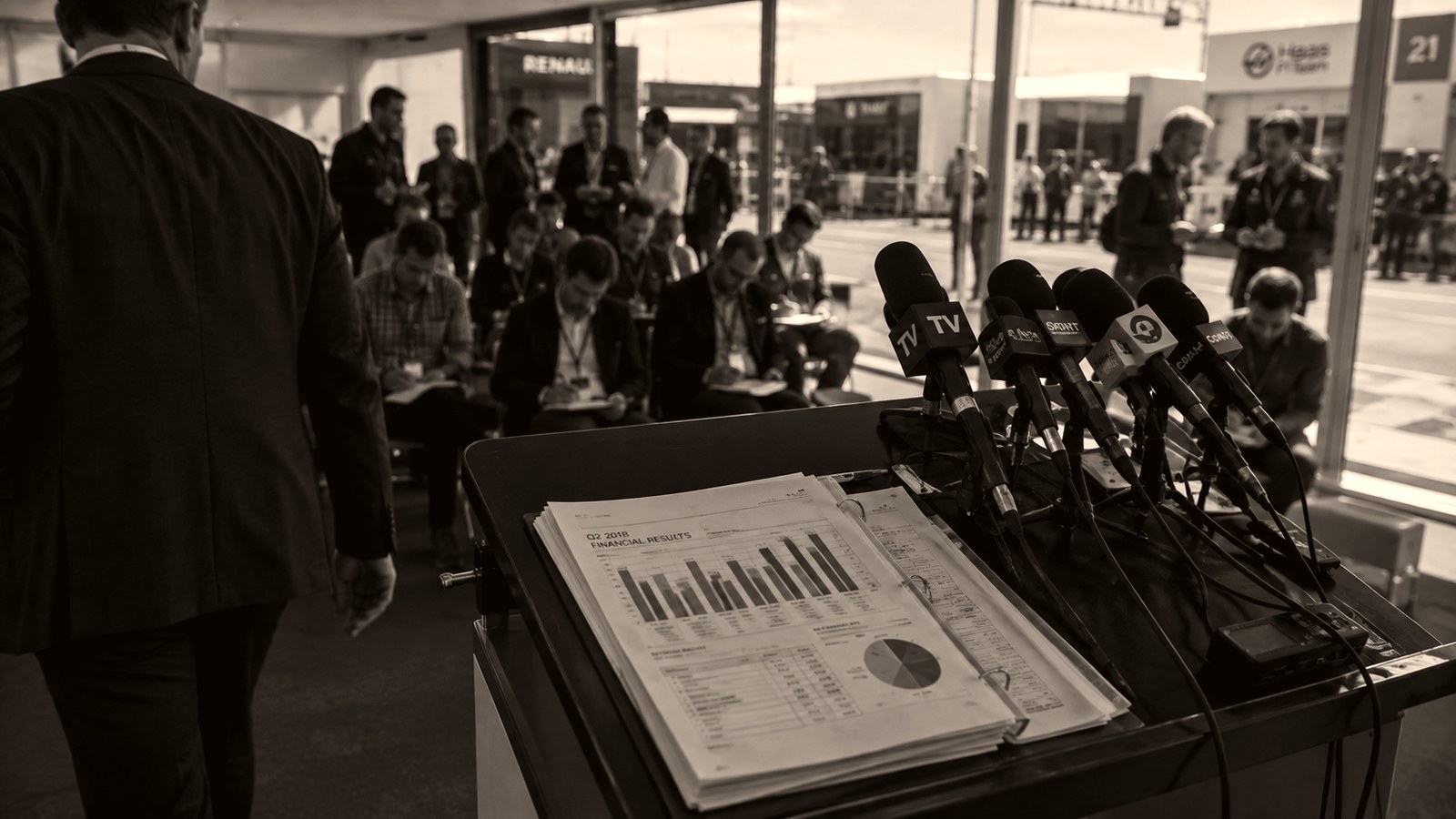 Atril de prensa en el paddock con un dossier de resultados financieros abierto y un bosque de micrófonos de televisión, mientras un directivo se aleja de espaldas y los periodistas toman notas
