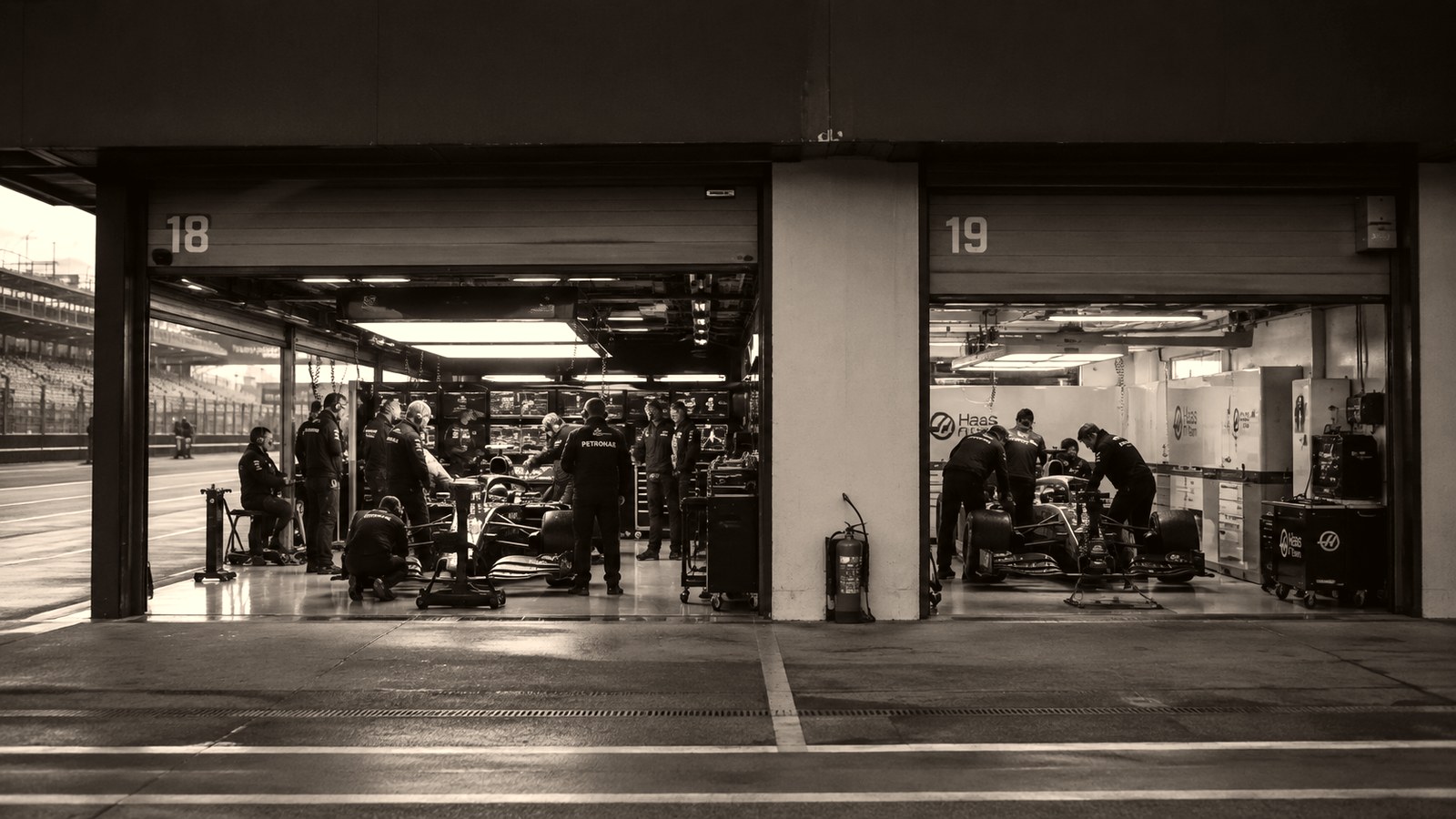 Dos garajes contiguos del pit lane, el 18 y el 19, iluminados de noche mientras los mecánicos trabajan sobre los monoplazas y el asfalto mojado refleja la luz